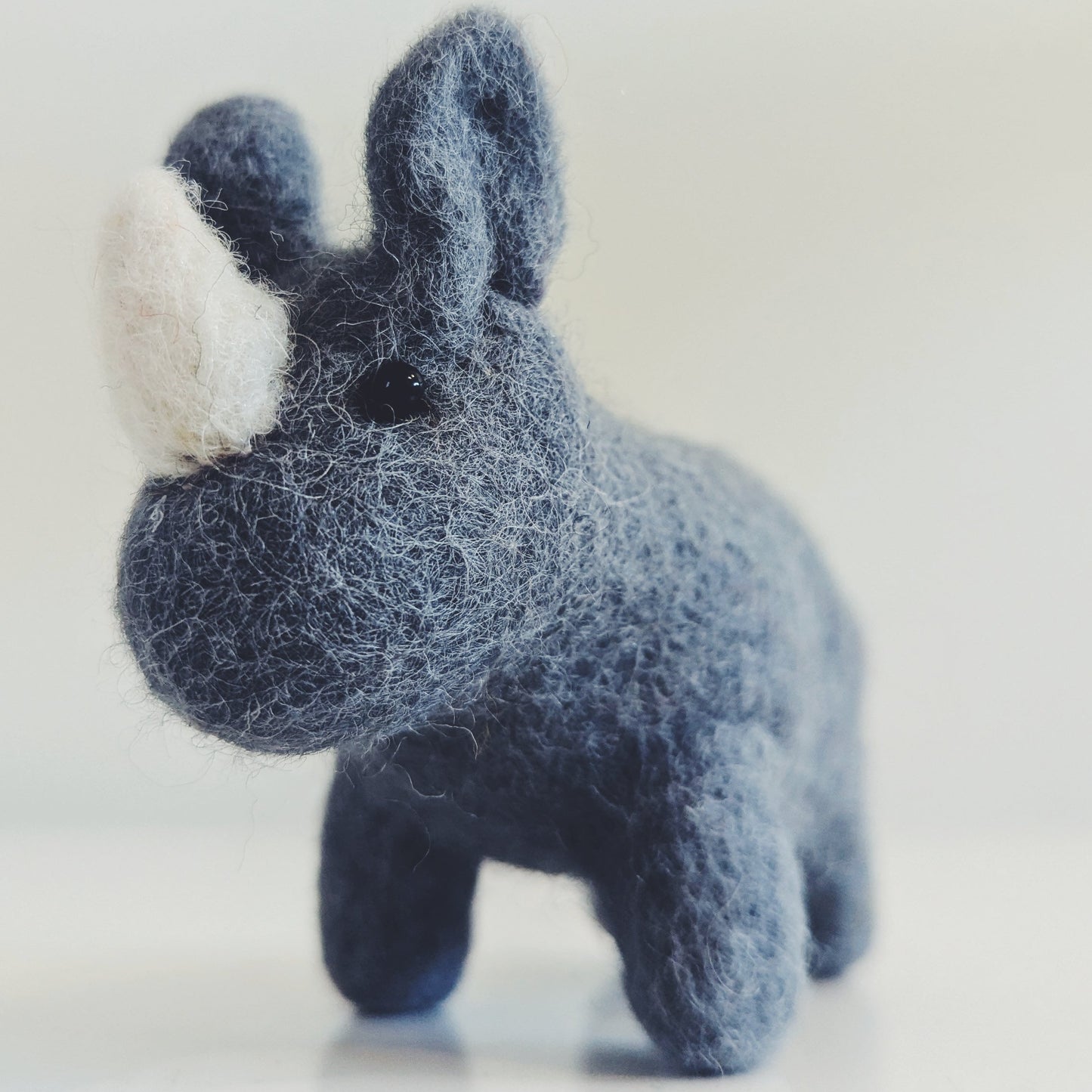 A small felted wool rhinoceros toy in dark blue-gray with a white horn, standing on a light gray surface against a pale gray background.