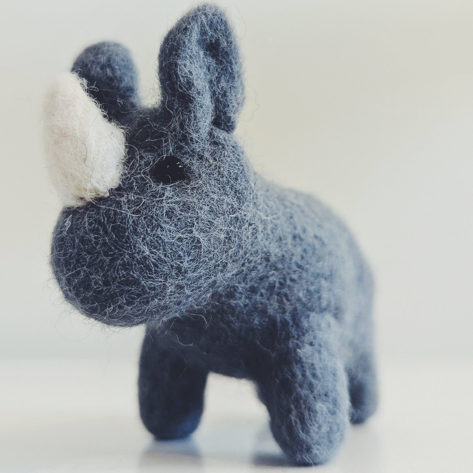 A small felted wool rhinoceros toy in dark blue-gray with a white horn, standing on a light gray surface against a pale gray background.