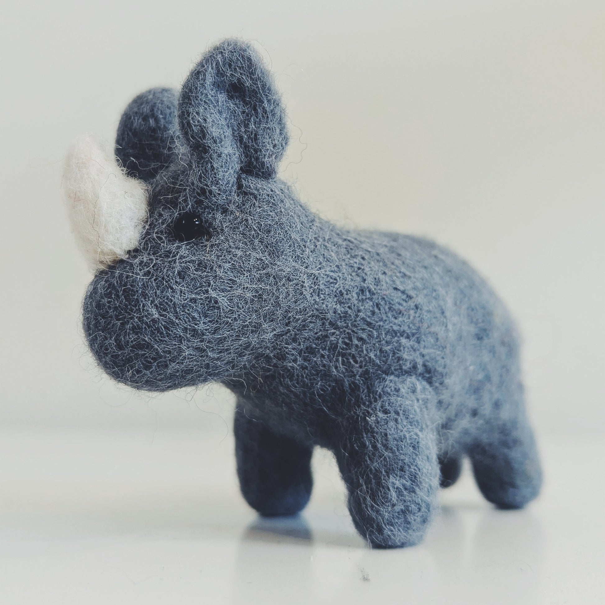 A small felt rhinoceros figurine in a textured slate blue color with a white horn, standing on a light reflective surface against a neutral light gray background.