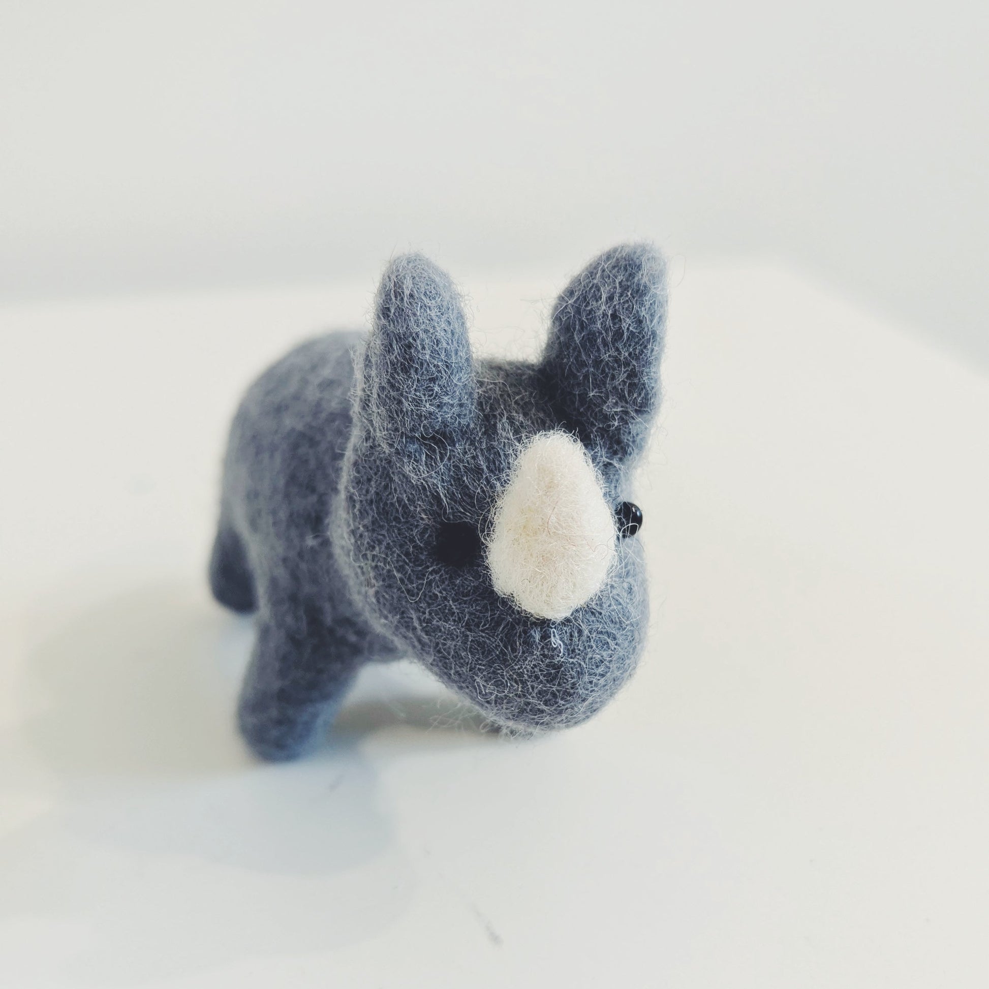 A small felted wool toy rhino, resembling a French bulldog, in a dark blue-gray color with a white snout, standing on a plain white surface.