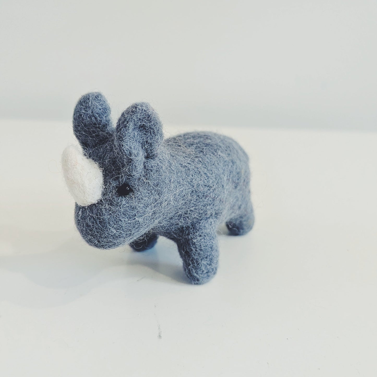 A small felt rhinoceros toy in a dusty blue-gray color with a white horn, standing on a plain white surface against a light gray background.