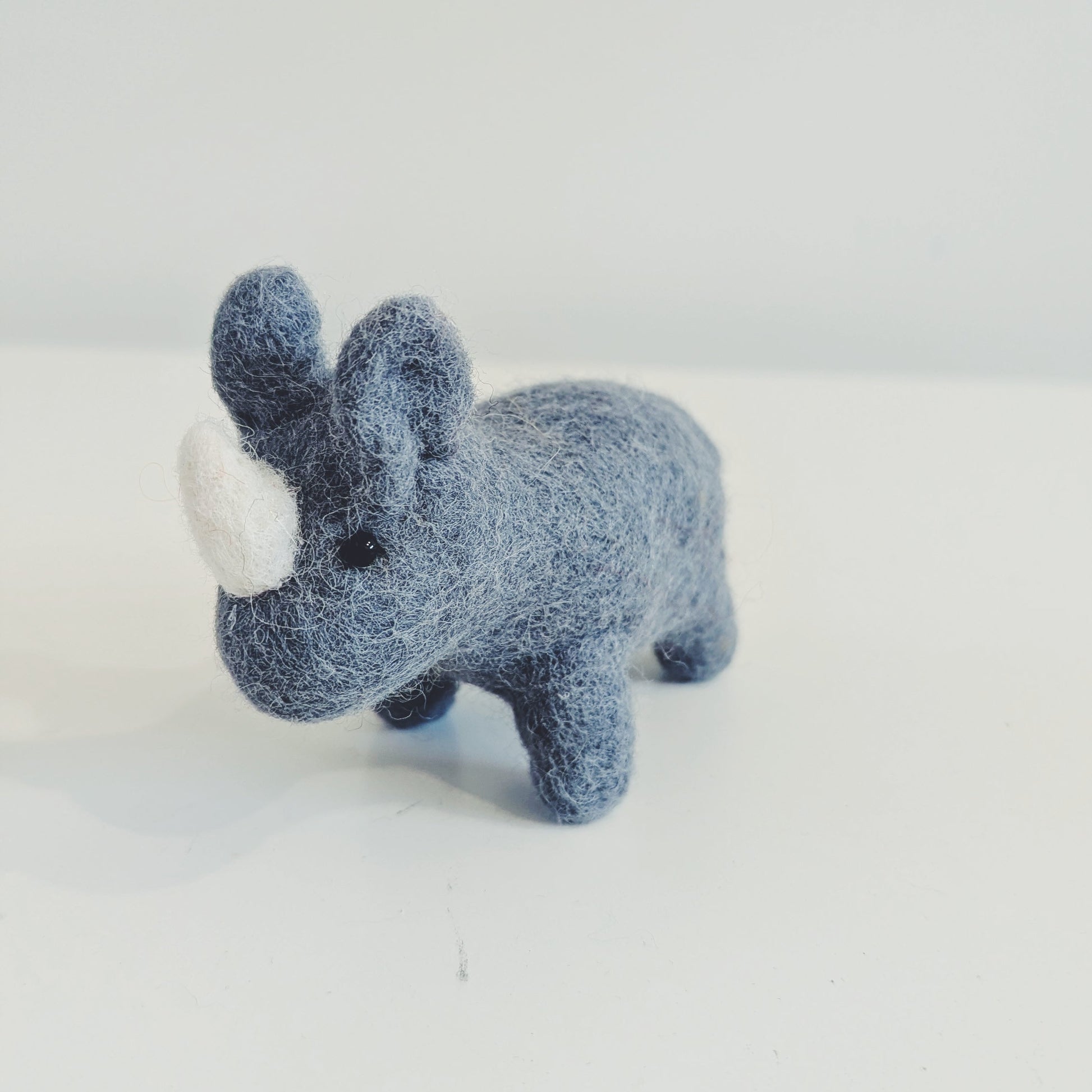 A small felt rhinoceros toy in a dusty blue-gray color with a white horn, standing on a plain white surface against a light gray background.
